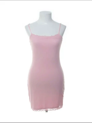 Rosa kort klänning med smala axelband - Söt rosa kort klänning med smala axelband och diskret volangkant vid halsringning och nederkant. Klänningen har en figurnära passform och är tillverkad i ett mjukt, stretchigt material som känns bekvämt mot huden. Perfekt för varma dagar eller som ett lagerplagg.