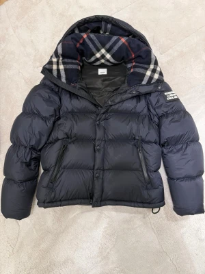 Marinblå pufferjacka från Burberry - Marin blå pufferjacka från Burberry med klassiskt rutigt foder i huvan och tydlig logotyp på ärmen. Jackan har dragkedjor på fickorna, knappar framtill och snörning i huvan. Perfekt för kalla dagar med sin tjocka och quiltade design. Storlek L