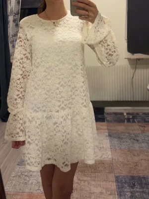 Vit spetsklänning med volangärm - Superfin vit kort klänning i spets med blommigt mönster. Klänningen har långa ärmar med volang vid ärmslut och en lös passform. Perfekt för sommarens alla tillfällen och har en romantisk vibe. Tillverkad i spetsmaterial som ger en luftig känsla. Helt oanvänd, prislappen sitter kvar, storlek Xs men passar som S också, perfekt till dig som tar studenten