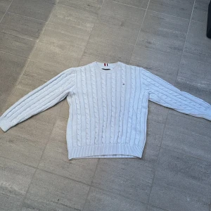Ljusblå kabelstickad tröja Tommy Hilfiger - Kabelstickad tröja i ljusblått från Tommy Hilfiger med rund halsringning och diskret logga på bröstet. Tröjan har ribbade muddar vid ärmslut och nederkant, perfekt för en stilren och klassisk look. 