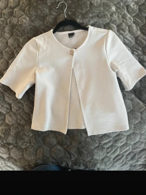 Vit kortärmad kofta med knapp - Stilren vit kofta med korta ärmar och rund halsringning. Koftan har en dekorativ metallknapp framtill och är gjord i ett mjukt, strukturerat tyg. Perfekt att slänga över en topp för en clean look.