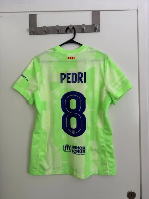 Barcelona matchtröja Pedri nr 8 - Snygg Barcelona matchtröja i ljusgrönt med tryck Pedri och nummer 8 på ryggen. Tröjan har klubbmärke, Nike-logga och Spotify som sponsor framtill. Tillverkad i lätt och ventilerande funktionsmaterial, perfekt för fotbollsträning eller match.