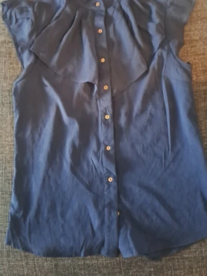 Mörkblå blus med volangdetalj - Snygg mörkblå blus med knappar framtill och volangdetalj över bröstet. Blusen är ärmlös och har en avslappnad passform, perfekt till jeans eller kjol. Tillverkad i ett lätt tyg som känns skönt mot huden.