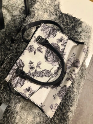 Vit handväska med blommönster - Snygg vit handväska från Fiorelli med svart blommönster och svarta handtag. Väskan har en strukturerad form och detaljer i silver. Perfekt för dig som gillar stilrena accessoarer med en twist.