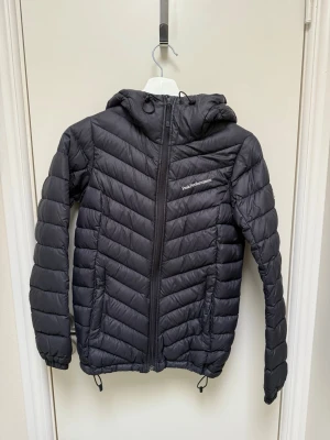 Svart dunjacka Peak Performance XS - Nypris: 2400kr Snygg svart dunjacka från Peak Performance med huva och dragkedja framtill. Jackan har en stilren design med diskret logga på bröstet och smidiga dragskor i nederkant. Perfekt för kalla dagar och enkel att matcha med olika outfits.