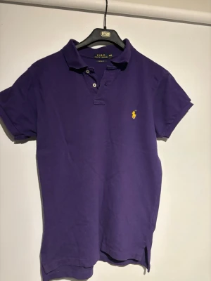 Lila piké från Ralph Lauren - Snygg lila pikétröja från Ralph Lauren med klassisk krage, korta ärmar och den ikoniska gula broderade loggan på bröstet. Stilren look. Sommar/vår plagg