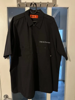 Svart Dream Theater skjorta - Svart skjorta med kort ärm från Dream Theater. Framsidan har två bröstfickor och broderad logga, baksidan har ett tryck med stadssilhuett och UFO. Klassisk krage och knäppning framtill. Perfekt för fans av bandet.