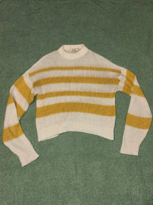 Randig stickad tröja  - Säljer en stickad tröja från NA-KD i vitt och senapsgult med breda horisontella ränder. Tröjan har en avslappnad passform, rund halsringning och långa ärmar. Materialet är mjukt och luftigt, perfekt för lager på lager under kyliga dagar.