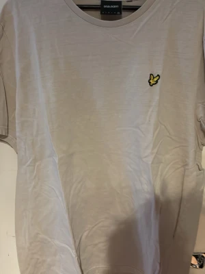 Beige t-shirt från Lyle & Scott - Snygg beige t-shirt från Lyle & Scott med klassisk rund hals och gul broderad logga på bröstet. Perfekt basic-plagg med stilren design som passar till det mesta. Tillverkad i mjuk bomull för skön känsla hela dagen.