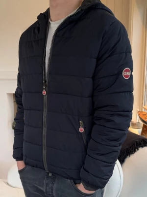 Svart pufferjacka från Colmar - Snygg svart pufferjacka från Colmar med huva och dragkedja framtill. Jackan har två fickor med dragkedja och Colmar-logga på ärmen. Perfekt för kalla dagar och ger en stilren look. Klassisk quiltad design som håller värmen.