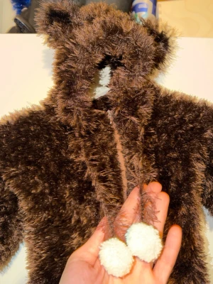 Brun fluffig björndräkt med huva - Supersöt brun björndräkt i mjukt, fluffigt material med huva och små öron. Dräkten har dragkedja framtill och vita bollar som dekoration i snörena. Perfekt för maskerad eller cosplay när du vill vara en gullig björn.