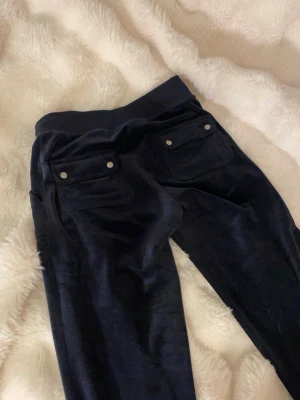 Mörkblå velourbyxor Juicy Couture - Mörkblå mjukisbyxor i velour från Juicy Couture med snören i midjan och stora fickor bak med silverfärgade knappar. Klassisk logga broderad på ena fickan fram. Perfekta för en chill och avslappnad stil. Säljer då de är försmå