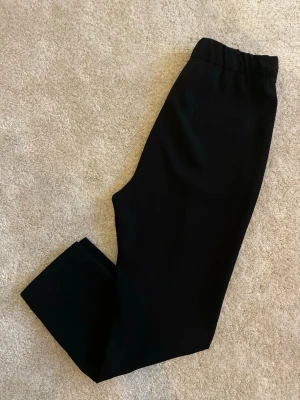 Svarta avslappnade byxor med snörning - Säljer ett par svarta kostymbyxor med avslappnad passform och elastisk midja med dragsko. Byxorna har raka ben och diskreta fickor på sidorna. Perfekta för en clean och stilren look.