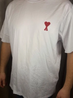 Vit t-shirt från Ami Paris med hjärtdetalj XL-XXL - Vit t-shirt från Ami Paris med röd broderad hjärtdetalj och bokstaven A på bröstet. Klassisk passform och rund halsringning. Tillverkad i mjuk bomull som känns skön mot huden. Perfekt för en clean och stilren look.