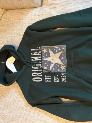 Mörkgrön hoodie från scndi - Snygg mörkgrön hoodie från scndi med huva och magficka. På bröstet finns ett stort tryck med texten 'ORIGINAL FIT EST. 2024' och en stjärna omgiven av mindre stjärnor. Perfekt för en avslappnad och cool stil. 