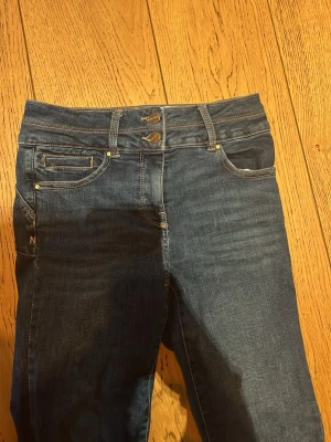 Blå bootcut jeans med dubbla knappar - Säljer ett par klassiska blå bootcut jeans med dubbla knappar i midjan och femficksmodell. Jeansen har en snygg tvätt och subtila slitningar framtill. Perfekta för dig som gillar en tidlös och avslappnad stil.