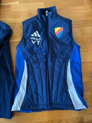 Blå dunväst från Adidas DIF - Sportig blå dunväst från Adidas med Djurgårdens IF-logga på bröstet. Västen har vita och grå detaljer på sidorna, hög krage och dragkedja framtill. Perfekt för dig som vill visa klubbkänsla och hålla värmen under träning eller på läktaren. Nypris 800kr. 