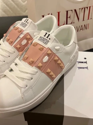 Valentino Rockstud sneakers vit/rosa - Säljer ett par Valentino Rockstud sneakers i vitt skinn med ljusrosa detaljer och ikoniska nitar. Skorna har snörning, rund tå och platt sula. Perfekta för dig som vill ha en lyxig och edgy touch till din outfit.