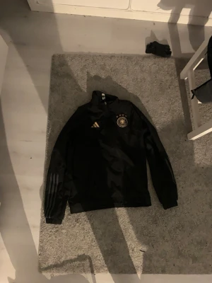 Svart Adidas Tyskland träningströja - Svart träningströja från Adidas med Tysklands landslagsmärke på bröstet och Adidas-logga i guld. Tröjan har dragkedja i halsen och ribbade muddar. Perfekt för fotbollsträning eller sportiga outfits.
