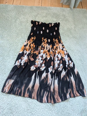 Blommig svart klänning med smock - Säljer en svart klänning med smock upptill och blommigt mönster i vitt, beige och orange. Funkar även som kjol. Väldigt stretchig så passar många storlekar, typ från XS till m/l. Köpt utomlands så ingen lapp. 
