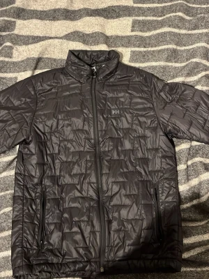Svart dunjacka från Helly Hansen - Snygg svart dunjacka från Helly Hansen med quiltat mönster och hög krage. Jackan har dragkedja framtill, två sidofickor med dragkedja och HH-logga på bröstet. Perfekt för kalla vinterdagar och riktigt stilren design.