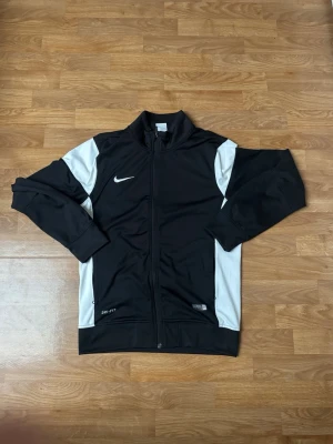 Nike tracksuit  - Nike träningsjacka i svart med vita paneler längs sidorna och ärmarna. Jackan har hel dragkedja, ståkrage och Nike-logga på bröstet samt bak i nacken. Tillverkad i ett lätt och stretchigt material, perfekt för träning eller uppvärmning.