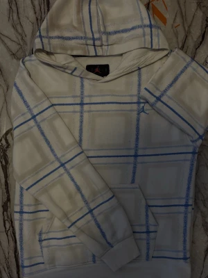 Vit hoodie med blått rutmönster - Snygg vit hoodie från Jordan med blått rutmönster och klassisk känguruficka. Hoodien har huva och ribbade muddar vid ärmslut och nederkant. Perfekt för en avslappnad och trendig stil.