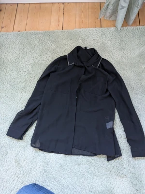 Svart skjorta med pärldetaljer - Stilren svart skjorta med lång ärm och pärldetaljer längs kragen. Skjortan har två bröstfickor och knäppning framtill. Tunt, lätt material som ger en luftig känsla. Perfekt för dig som gillar en enkel men ändå unik look.