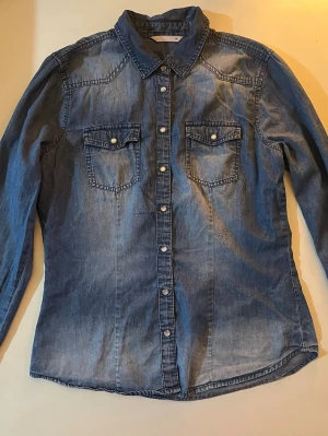 Blå jeansskjorta från ONLY - Snygg blå jeansskjorta från ONLY med klassisk krage, två bröstfickor med knappar och långa ärmar. Skjortan har tryckknappar framtill och är tillverkad i mjuk bomull. Perfekt för en avslappnad och trendig look.