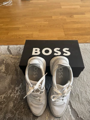 Vita sneakers från BOSS - Snygga vita sneakers från BOSS med svarta och grå detaljer. Skorna har en sportig design med grov sula och klassisk snörning. Tillverkade i en mix av läder och textil för en modern look. Perfekta för dig som gillar stilrena och trendiga sneakers.