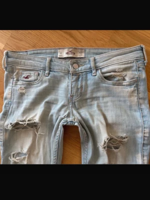 Ljusblå slitna jeans från Hollister - Säljer ett par ljusblå jeans från Hollister med trendiga slitningar och hål på både lår och knän. Jeansen har en klassisk femficksmodell och raka ben. Perfekta för en avslappnad och cool stil.