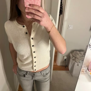 Vit kortärmad kofta med knappar - Säljer en söt cremevit kofta med korta ärmar och svarta knappar framtill. Koftan har rund halsringning och är stickad i ett mjukt material. Perfekt att styla med jeans eller kjol för en trendig look. Använd fåtal gånger. Är öppen för prisförslag 