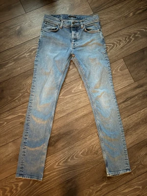  Nudie grim tim - Säljer ett par ljusblå Nudie grim Tim Jeans med klassisk femficksdesign och slitna detaljer. Jeansen har en smal passform och är tillverkade i bomull med stretch för extra komfort. Perfekta för en avslappnad och trendig look.