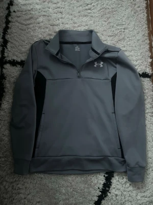Grå halvzip från Under Armour - Snygg grå halvzip-tröja från Under Armour med svart logga på bröstet. Tröjan har hög krage, dragkedja framtill och svarta paneler på sidorna. Tillverkad i ett mjukt och stretchigt material som passar perfekt till lager-på-lager.