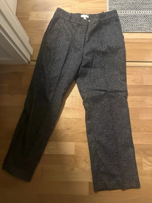 Svartmelerade kostymbyxor från H&M - Snygga  kostymbyxor från H&M i klassisk rak modell. Byxorna har diskreta fickor i sidorna och hällor för bälte. Materialet har en lätt melerad struktur som ger en stilren look. Perfekta för dig som vill ha en clean och dressad stil.