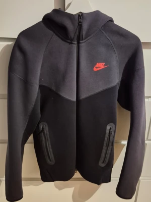 Svart Nike tech fleece hoodie med dragkedja - Snygg svart hoodie från Nike tech med röd logga på bröstet. Tröjan har hel dragkedja framtill, två fickor med dragkedja och en stor huva. Tillverkad i mjukt och stretchigt material som passar perfekt till en sportig stil.