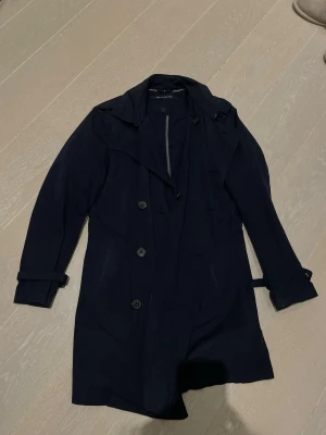 Mörkblå trenchcoat från Marc O'Polo - Stilren mörkblå trenchcoat från Marc O'Polo med klassisk dubbelknäppning, bälte i midjan och slag vid kragen. Jackan har långa ärmar med justerbara remmar vid ärmsluten och är tillverkad i ett lätt tyg som passar perfekt för säsongsskiftet.
