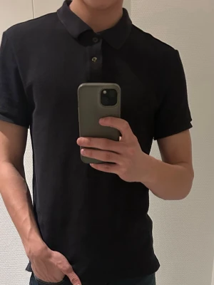 Svart slim fit pikétröja från H&M - Säljer en svart pikétröja från H&M i slim fit-modell. Tröjan har en krage, korta ärmar och två knappar framtill. Personligt perfekt för en clean och enkel look. 