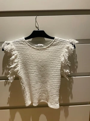 Vit topp med spetsvolang från Zara - Superfin vit topp från Zara med korta ärmar och volangdetaljer i spets längs axlarna. Stickad struktur 