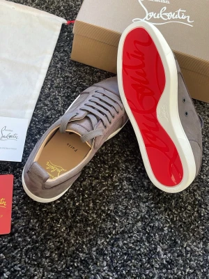 Gråa Christian Louboutin sneakers - Snygga gråa sneakers från Christian Louboutin med klassisk röd sula och diskret logga på insidan. Skorna är låga och har ovandel i mocka med matchande grå skosnören. Perfekta för dig som vill sticka ut med exklusiv stil. Helt nya aldrig använda 10/10 skick. Allt ingår. Snabb affär bättre pris
