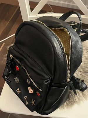 Svart ryggsäck med pins och nitar - Cool svart ryggsäck i skinnimitation med guldfärgade dragkedjor och prickigt innerfoder. Framsidan är dekorerad med olika pins och nitar i form av stjärnor, bokstäver och hjärtan. En pin fattas men syns inte så mycket. Justerbara axelremmar och praktiskt ytterfack med dragkedja.