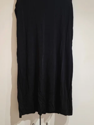 Svart långklänning från Indiska - En enkel och stilren svart långklänning från Indiska. Klänningen har rak passform och rund halsringning. Perfekt basplagg att styla på olika sätt. Materialet känns mjukt och följsamt.