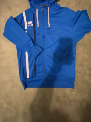 Blå hoodie från Erreà med dragkedja - Snygg blå hoodie från Erreà med hel dragkedja framtill, vita och mörkblå ränder på vänster sida samt vita snören i huvan. Tillverkad i ett mjukt och sportigt material, perfekt för en avslappnad stil. Logga på bröstet och ribbade muddar vid ärmslut och nederkant.