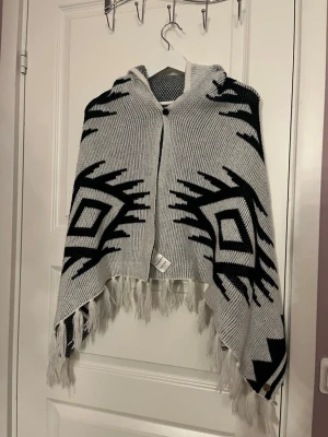 Poncho med huva från KappAhl - Snygg stickad poncho från KappAhl i grått och svart med grafiskt mönster. Ponchon har huva, fransar längs nederkanten och knäppning framtill. 