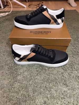 Burberry sneakers med rutigt mönster - Snygga svarta sneakers från Burberry med klassiskt beige, svart och vitt rutmönster på sidorna. Skorna har svarta skosnören, vit sula och detaljer i mocka och textil. Perfekta för dig som vill ha en stilren och trendig look.