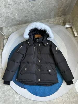 Svart dunjacka med pälskantad huva - Snygg svart dunjacka från Moose Knuckles med stor huva kantad av vit fuskpäls. Jackan har flera fickor med tryckknappar, dragkedja framtill och logga på ärmen. Perfekt för kalla vinterdagar och riktigt varm.