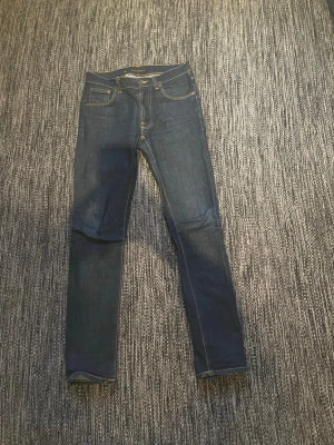 Mörkblå skinny jeans från Nudie Jeans - Säljer ett par mörkblå skinny jeans från Nudie Jeans med klassisk femficksdesign och kontrastsömmar. Jeansen har en smal passform och är tillverkade i slitstark denim av bomull. Perfekta för dig som gillar en clean och tidlös look. Var inte rädd för att skicka om du har någon fråga eller vill ha fler bilder!