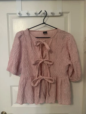 Blus med rosetter  - Supersöt ljusrosa blus från Gina Tricot med strukturmönster och tre dekorativa knyt framtill. Blusen har rund halsringning, volang nedtill och halvlånga puffärmar. 💓