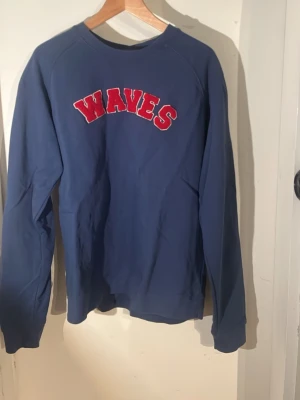 Blå sweatshirt med röd WAVES-text - Säljer en blå sweatshirt med röd text 'WAVES' i varsity-stil på bröstet. Tröjan har rund halsringning och långa ärmar. Perfekt för dig som gillar en sportig och avslappnad look. Materialet känns mjukt och bekvämt, troligen bomull eller bomullsblandning.