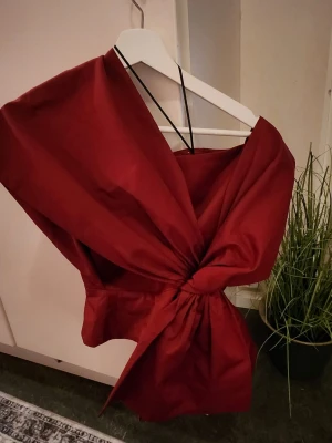 Röd omlott topp med stor rosett - Supersnygg röd omlott topp med en stor rosett framtill och tunna axelband. Toppen har en unik design som verkligen sticker ut och passar perfekt till dig som vill ha något extra. Materialet känns som bomull eller polyester.